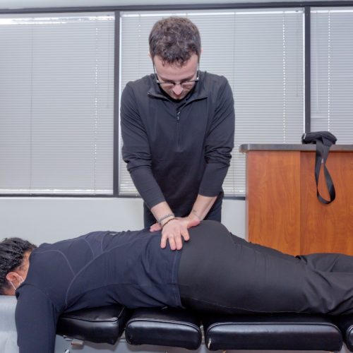 Home 5 Male chiropractor performing hands-on spinal adjustment on patient lying face down on treatment table.